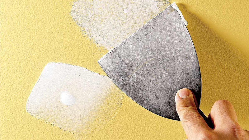 image of yellow wall with two holes that have been repaired, hand holding putty knife